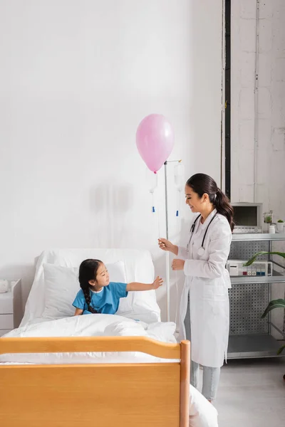 Sorrindo médico no branco casaco de pé com festivo balão perto asiático menina no hospital cama — Fotografia de Stock