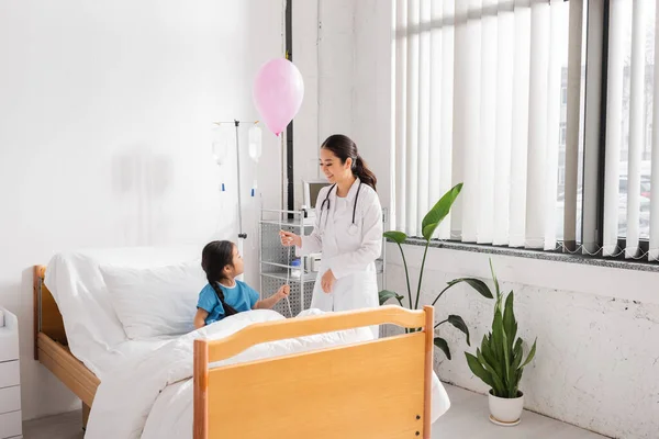 Smiling asian doctor holding festive balloon near girl sitting on bed in modern hospital ward — Stock Photo