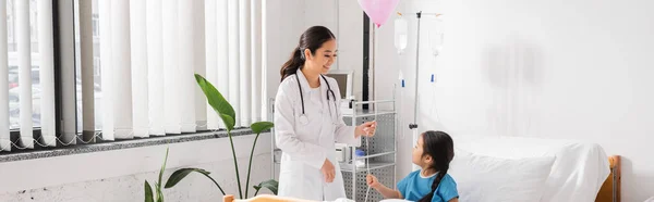 Happy asian doctor in white coat holding festive balloon near kid in modern pediatric clinic, banner — Stock Photo