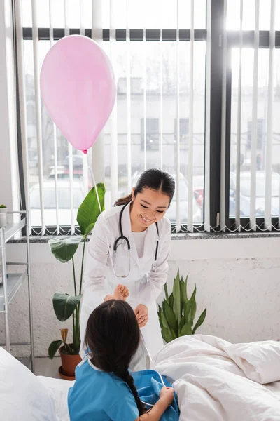 Alegre asiático médico dando festivo globo a poco paciente en pediátrico hospital - foto de stock