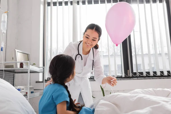 Young asian pediatrician with festive balloon smiling near girl sitting on bed in hospital ward — Stock Photo