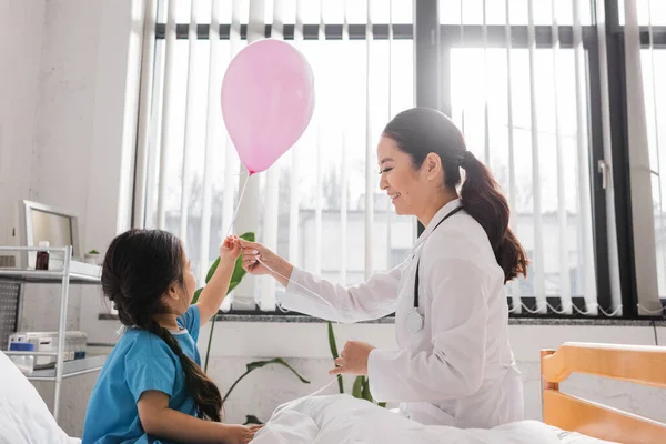 Vista laterale di felice asiatico medico holding festive palloncino vicino a bambina in ospedale reparto — Foto stock