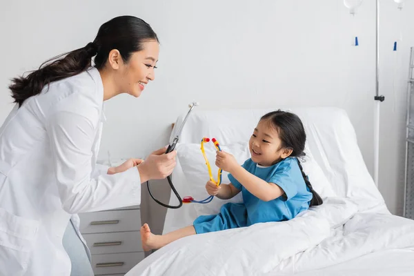 Side view of cheerful asian girl showing toy stethoscope to smiling doctor in hospital ward — Stock Photo