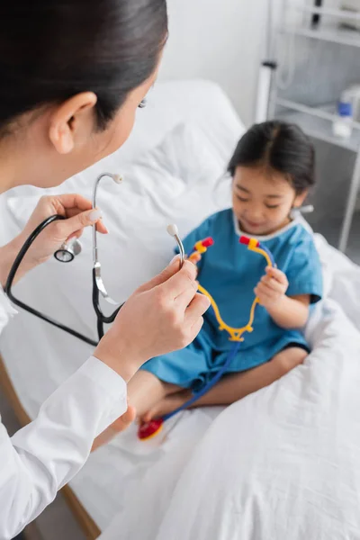 Молодий лікар біля розмитої азіатської дівчини тримає іграшковий стетоскоп, сидячи на лікарняному ліжку — стокове фото