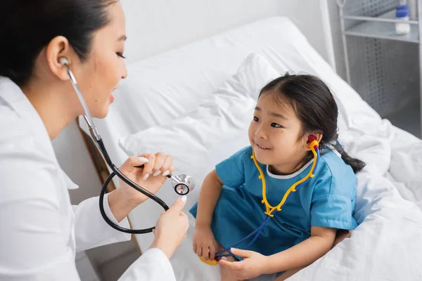 Pediatrician showing stethoscope to smiling asian girl sitting on bed in hospital ward — Stock Photo