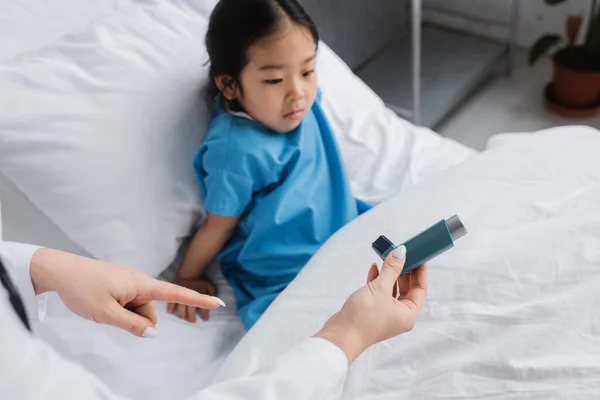 Médico señalando inhalador cerca desanimado asiático chica sentado en hospital cama - foto de stock