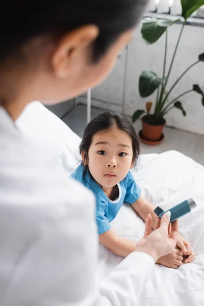 Blurred pediatrician showing inhaler to surprised asian child in hospital ward — Stock Photo