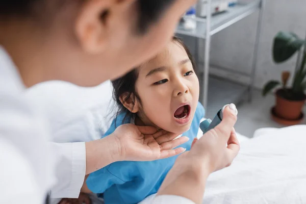 Хвора азіатська дівчина відкриває рот біля розмитого педіатра з інгалятором у лікарняному палаті — стокове фото