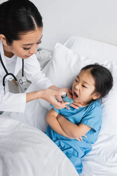Joven pediatra celebración inhalador cerca asiático chica con abierto boca y brazos cruzados en hospital cama - foto de stock