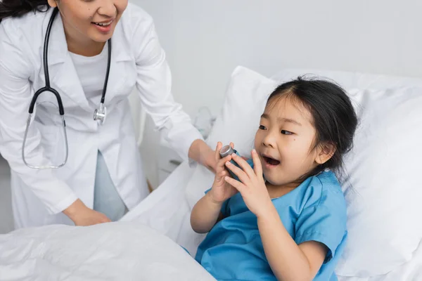 Маленькая азиатская девочка с открытым ртом держит ингалятор рядом с доктором в педиатрической клинике — стоковое фото
