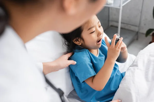 Pediatra borrosa tocando hombro de niño asiático usando inhalador en sala de hospital - foto de stock
