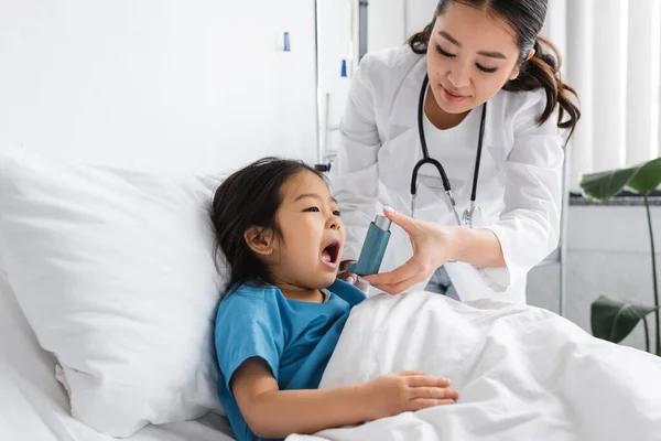Jeune asiatique pédiatre tenant inhalateur près de peu patient couché avec la bouche ouverte sur lit d'hôpital — Photo de stock