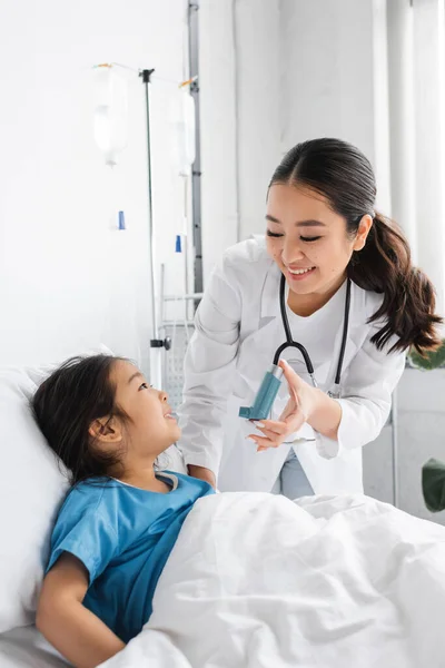 Усміхнений лікар тримає інгалятор біля позитивної дівчини на ліжку в лікарняному палаті — стокове фото