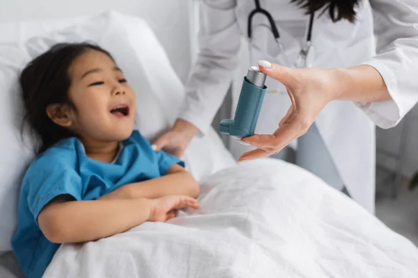 Verschwommener Kinderarzt hält Inhalator in der Nähe von asiatischem Kind, das Mund auf Krankenhausbett öffnet — Stockfoto