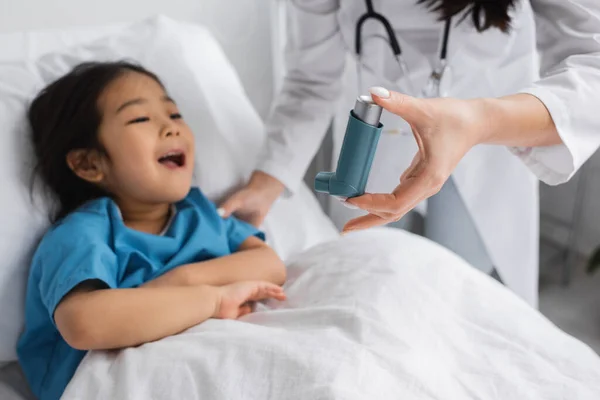 Blurred pediatrician holding inhaler near asian child opening mouth on hospital bed — Stock Photo