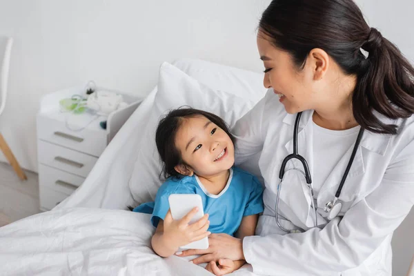 Cheerful asian girl looking at young pediatrician and taking selfie on mobile phone in clinic — Stock Photo
