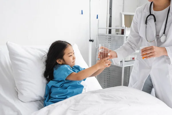 Médico no casaco branco segurando pílulas recipiente e dando vidro de água para menina asiática na cama do hospital — Fotografia de Stock