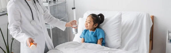 Doctor in white coat holding medication and glass of water near asian girl smiling on hospital bed, banner — Stock Photo