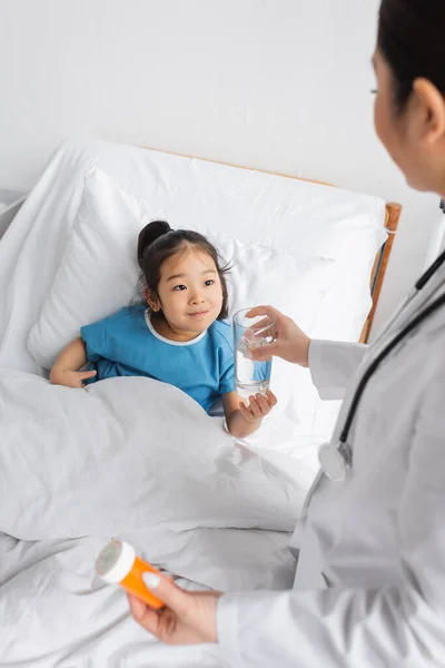 Весела азіатська дитина бере склянку води від лікаря, тримаючи тару для таблеток у лікарняному палаті — стокове фото