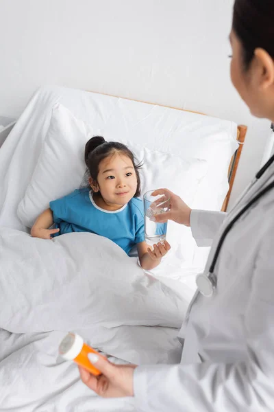Cheerful asian child taking glass of water from doctor holding pills container in hospital ward — Stock Photo