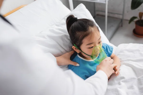 Borrosa médico ajuste de oxígeno máscara en enfermo asiático niño en hospital cama - foto de stock