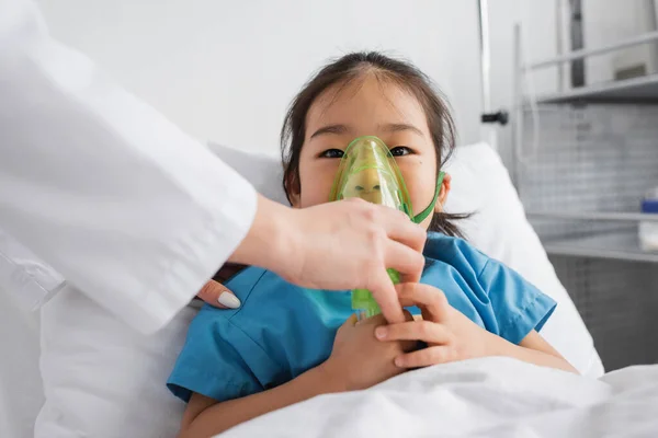 Pequeño asiático chica respiración en oxígeno máscara cerca médico en hospital sala - foto de stock