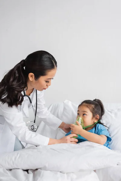 Young doctor helping asian girl breathing in oxygen mask on bed in hospital ward — Stock Photo