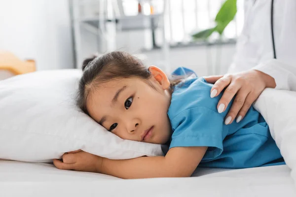 Doctor calming upset asian girl lying on bed in hospital ward — Stock Photo