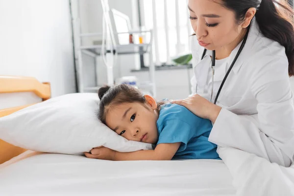 Young asian doctor calming sad child lying on bed in clinic — Stock Photo