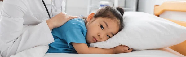 Enfermo y frustrado asiático niño mirando a cámara mientras se encuentra en cama de hospital cerca de médico, pancarta - foto de stock