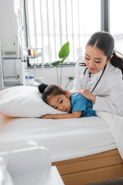 Joven pediatra calmando enfermo y triste asiático chica acostado en cama en hospital - foto de stock
