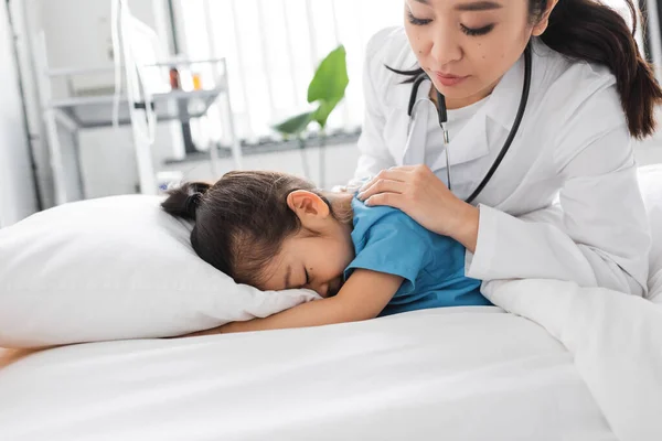 Asian doctor calming depressed child obscuring face with pillow on hospital bed — Stock Photo