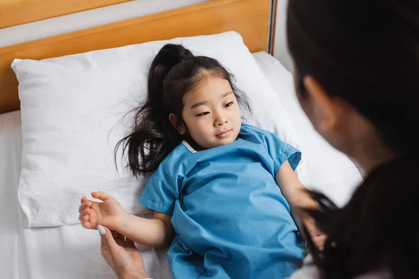 Высокий угол обзора маленькой азиатской девочки, лежащей на больничной койке рядом с размытым врачом — стоковое фото