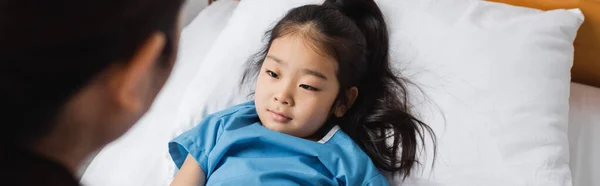 Diseased asian girl looking at blurred pediatrician while lying on hospital bed, banner — Stock Photo