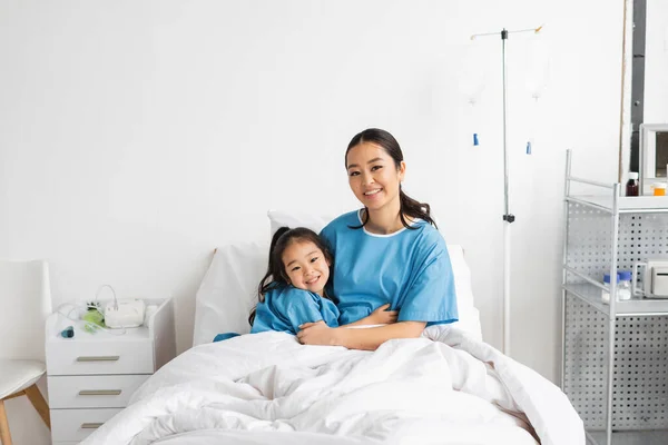 Fröhliche asiatische Mutter und Kind umarmen sich und schauen in die Kamera auf dem Krankenhausbett — Stockfoto