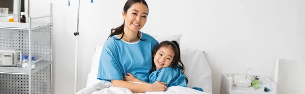 Joyful asian mother and daughter smiling at camera on bed in hospital ward, banner — Stock Photo
