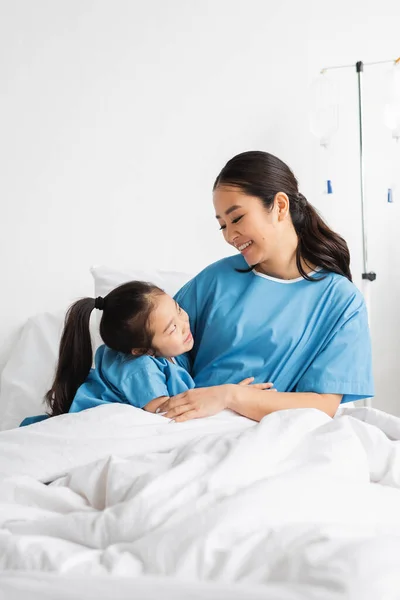Щаслива азіатська мати і дочка обіймаються і дивляться один на одного на лікарняному ліжку — стокове фото