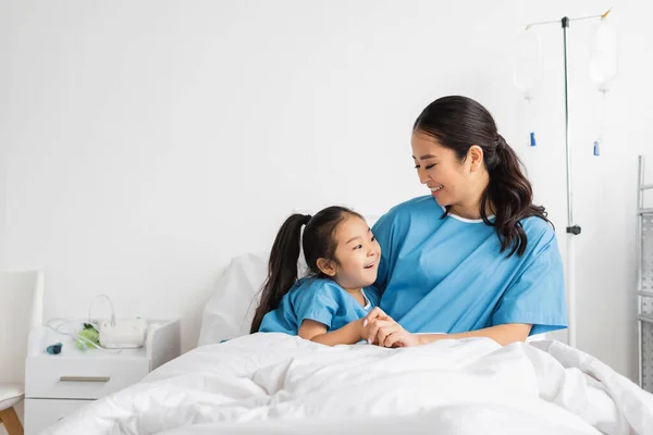 Young asian woman holding hands of smiling daughter on bed in hospital ward — Stock Photo