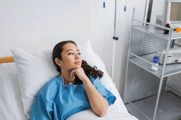 Mujer asiática soñadora sosteniendo la mano cerca de la barbilla y mirando hacia otro lado en la cama en el hospital - foto de stock