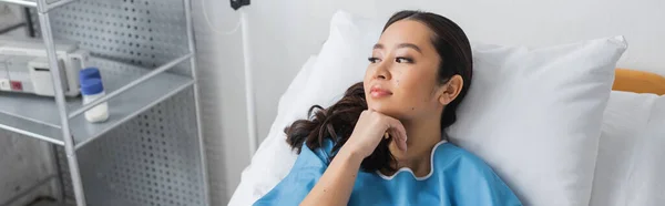 Mujer asiática soñadora y sonriente cogida de la mano cerca de la barbilla y mirando hacia otro lado en la cama del hospital, pancarta - foto de stock