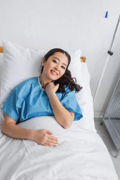 Высокий угол обзора довольной азиатской женщины, смотрящей в камеру, лежащей на кровати в палате больницы — стоковое фото