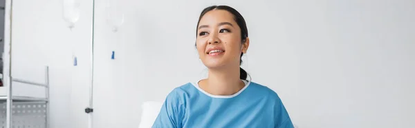 Молодая азиатка в больничном платье улыбается и смотрит в сторону в клинике, баннер — стоковое фото