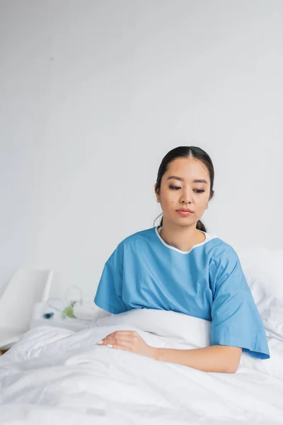 Depressed asian woman in hospital gown thinking while sitting on bed in clinic — Stock Photo