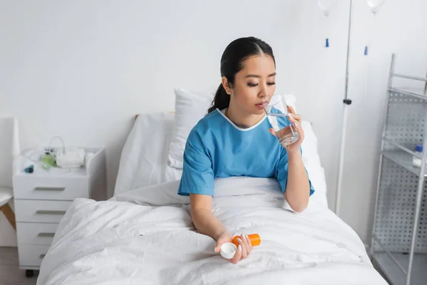 Joven asiático mujer beber agua y celebración píldoras contenedor mientras sentado en cama en hospital sala - foto de stock