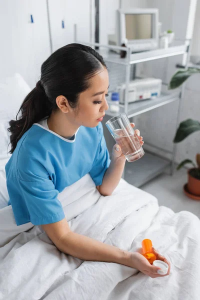 Brünette asiatische Frau im Krankenhauskleid sitzt auf dem Bett mit Glas Wasser und Pillen Behälter — Stockfoto