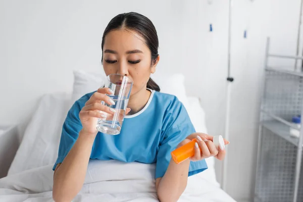 Jovem asiático mulher beber água enquanto segurando pílulas recipiente na cama na clínica — Fotografia de Stock