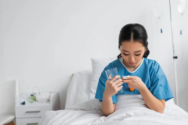 Junge Asiatin schaut in Tablettenbehälter, während sie ein Glas Wasser auf dem Krankenhausbett hält — Stockfoto