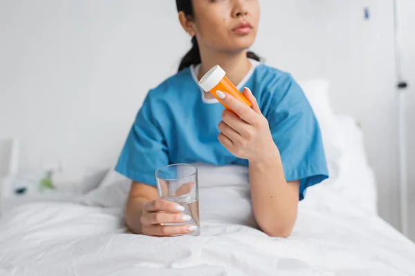Vue partielle de la femme en robe d'hôpital tenant l'eau et récipient de pilules sur le lit dans la clinique — Photo de stock