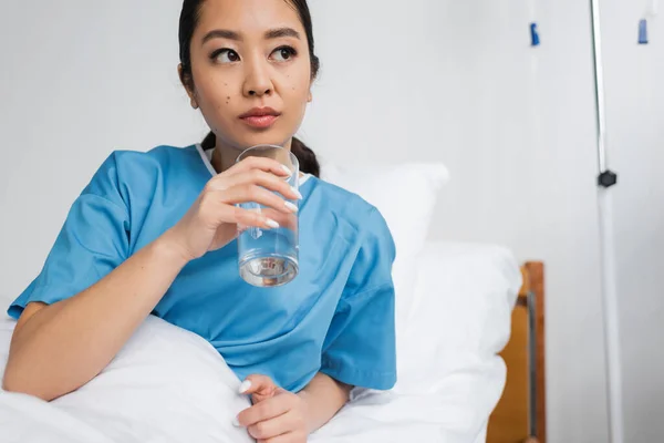 Pensativa mujer asiática sosteniendo vaso de agua y mirando hacia otro lado en hospital sala - foto de stock