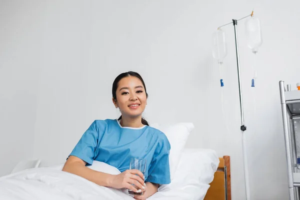 Cheerful asian woman holding glass of water and looking at camera on hospital bed — Stock Photo